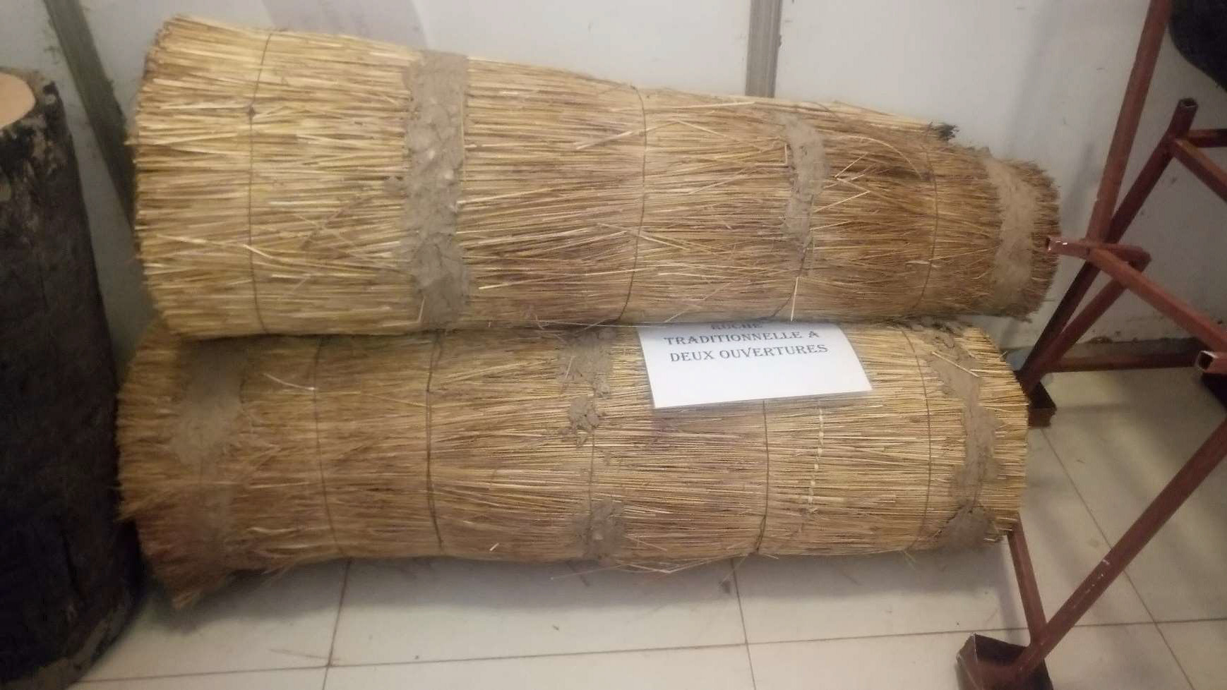 Two traditional beehives, crafted from bound straw and secured with natural ropes, are stacked on the ground. A sign reading "Traditionnelle à deux ouvertures" indicates their design. In the background, parts of a metal frame and a wall are visible.