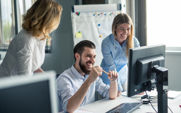 Ein Mann sitzt bei Tisch, lacht und schaut auf den Computer. Links und rechts von ihm sind zwei Frauen, die ebenfalls lachen und auf den Computerbildschirm schauen. 