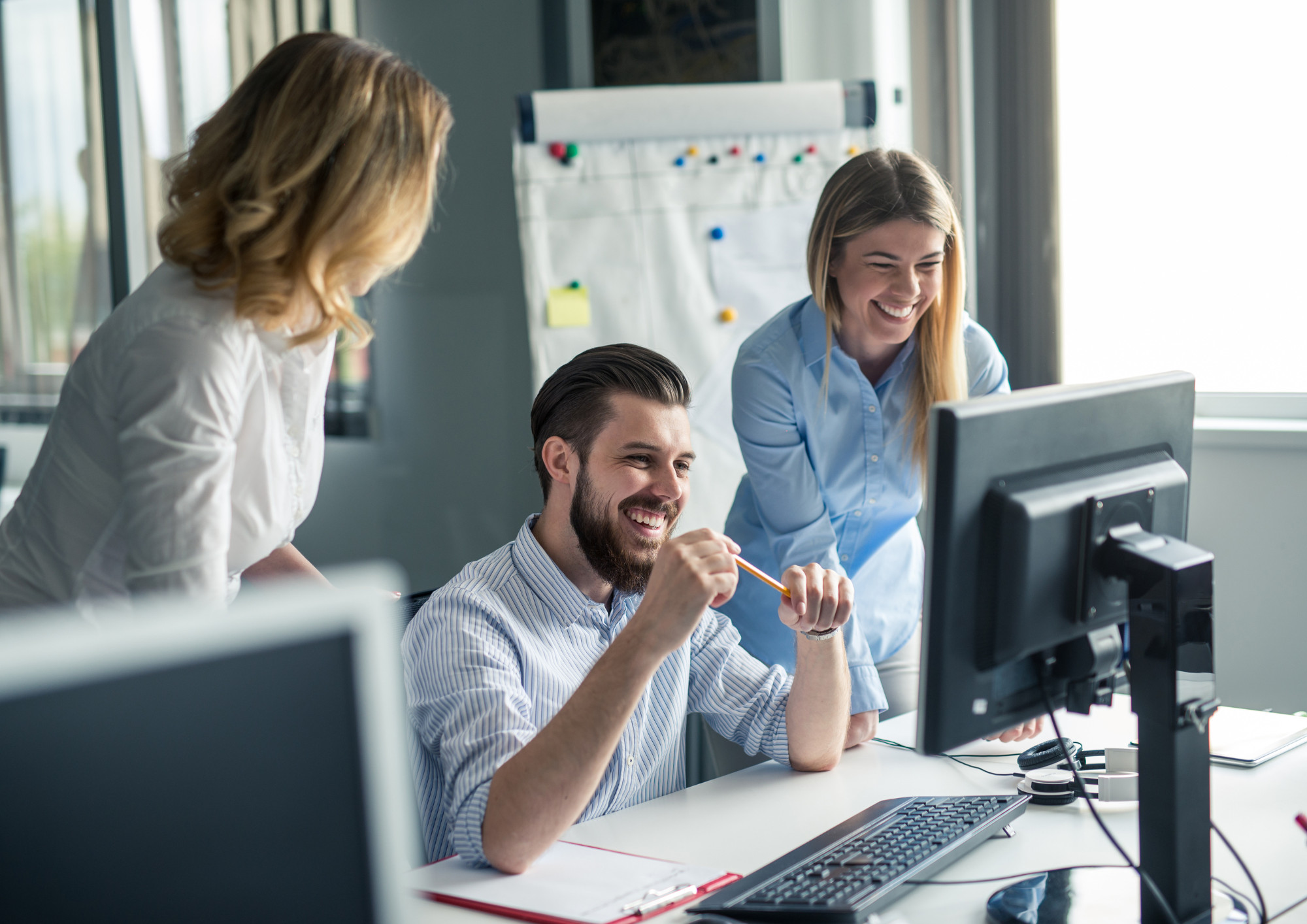 Ein Mann sitzt bei Tisch, lacht und schaut auf den Computer. Links und rechts von ihm sind zwei Frauen, die ebenfalls lachen und auf den Computerbildschirm schauen. 